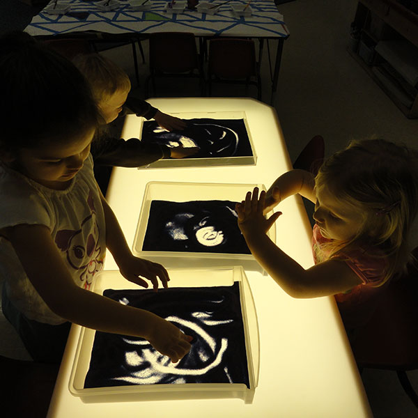 Kids playing with a box of sand on a lighted table.